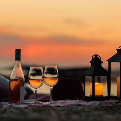 Summer sea sunset. Romantic picnic on the beach. Bottle of wine, glasses, candles, plaid and pillows. Selective focus.